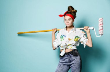 Beyaz tişörtlü, boyalı ve kot pantolonlu genç cesur kız omuzunda boya silindiri taşıyor.