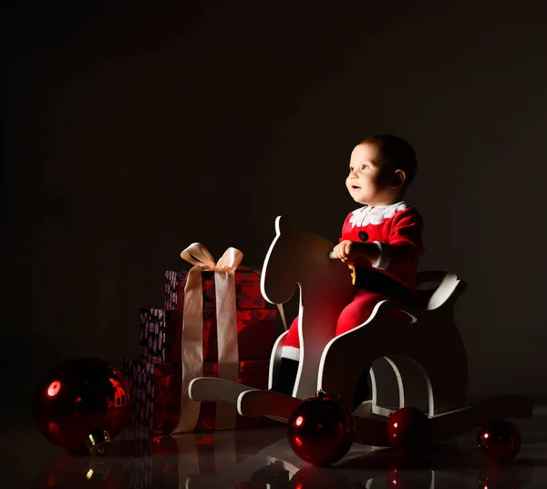 Mały chłopiec w stroju Świętego Mikołaja i kapelusz jeździ drewnianym koniem dla dzieci huśtawka z pudełkami noworocznych prezentów — Zdjęcie stockowe