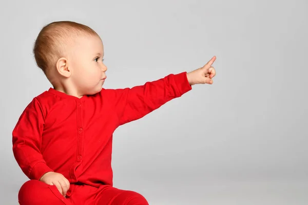 Pequeño niño apunta con el dedo hacia arriba lado en traje de bebé rojo con espacio de texto libre en gris — Foto de Stock