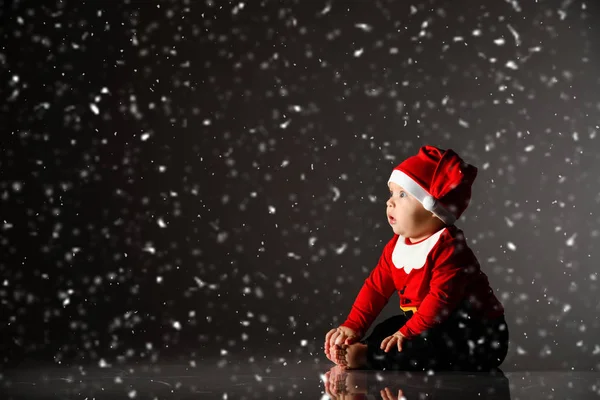 Χαμογελώντας βρέφος αγοράκι νήπιο σε κόκκινο καπέλο Χριστούγεννα και το νέο έτος κοστούμι κάθεται στον πάγο κάτω από το χιόνι κοιτάζοντας πάνω — Φωτογραφία Αρχείου
