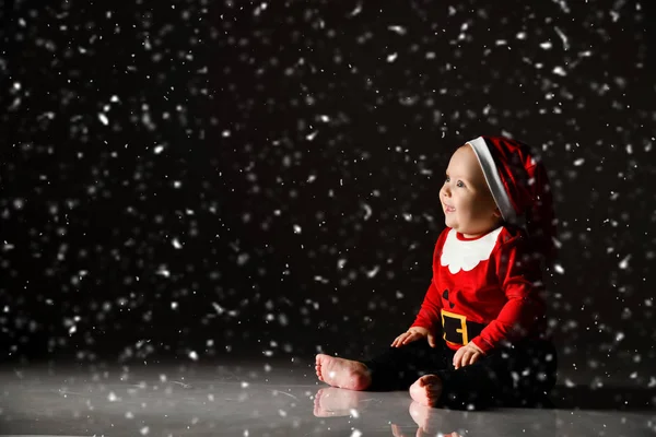 Χαμογελώντας βρέφος αγοράκι νήπιο σε κόκκινο καπέλο Χριστούγεννα και το νέο έτος κοστούμι κάθεται στον πάγο κάτω από το χιόνι κοιτάζοντας πάνω — Φωτογραφία Αρχείου