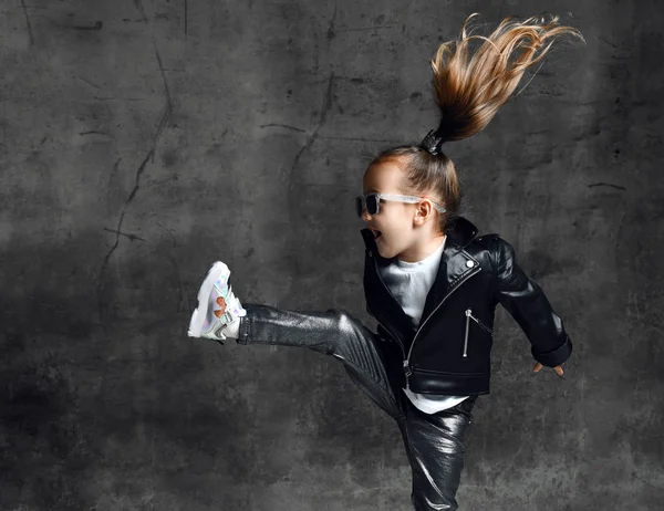 Ενεργό frolic κορίτσι παιδί με ασημένια κορώνα hairpin και σε δερμάτινο μπουφάν και γυαλιά ηλίου πηδά ανύψωση πόδι της ψηλά — Φωτογραφία Αρχείου