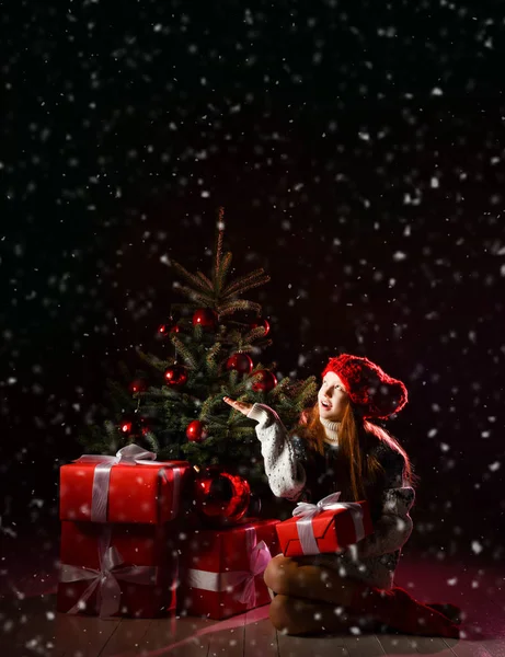 Niña sentada celebrando con abeto de Navidad de invierno decorado en cabeza roja tocando copos de nieve bajo la nieve — Foto de Stock