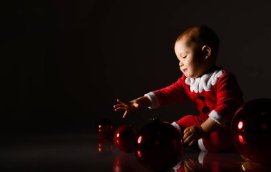 Gülümseyen bebek Noel Baba kostümlü bebek yerde yuvarlanan kırmızı cam toplarla oynuyor. 