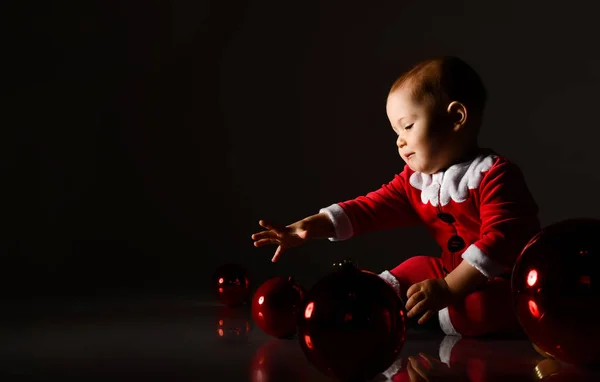 Bébé garçon souriant tout-petit en costume de Père Noël jouant avec des boules de verre rouge arbre de Noël roulant sur le sol — Photo