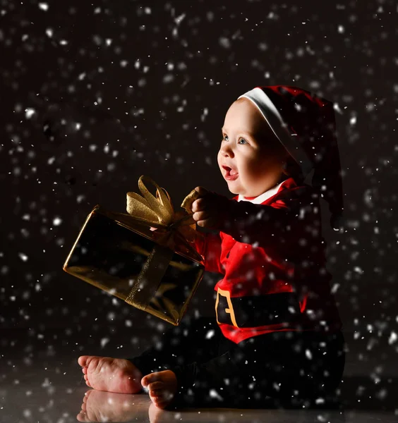 Lykkelig leende baby gutt sitter i juletåa kostyme holder julegave gave under snøen – stockfoto