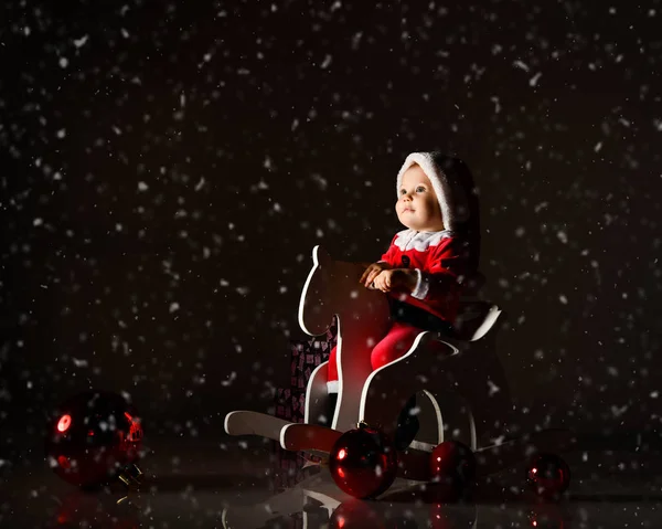 Enfant garçon tout-petit en costume de Père Noël claus et casquette de Noël monte son cheval en bois, regardant admirablement la chute de neige — Photo