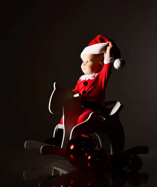 Mały chłopiec maluch w stroju Świętego Mikołaja jeździ na drewnianym koniu, patrząc w przyszłość, zdejmując świąteczną czapkę — Zdjęcie stockowe