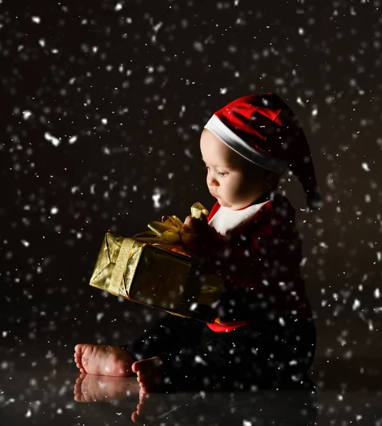 Szczęśliwy śmiech niemowlę chłopiec siedzi w stroju Mikołaja trzymając otwierające pudełko świąteczne pod śniegiem — Zdjęcie stockowe