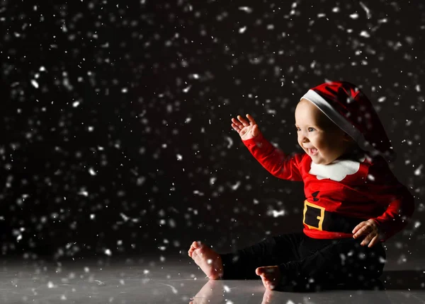 Szczęśliwy śmiech niemowlę chłopiec maluch siedzi w stroju Mikołaja i Boże Narodzenie czapka macha pod śniegiem na ciemno — Zdjęcie stockowe