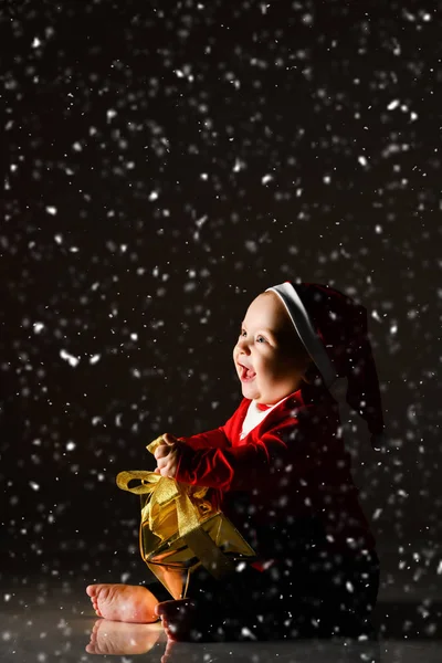 Niemowlę dziecko chłopiec dzieciak z niebieskimi oczami szczęśliwy uśmiech siedzi w kapeluszu Mikołaja ze złotym pudełku prezentów świątecznych — Zdjęcie stockowe