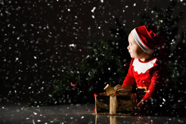 파란 눈을 가진 아기 아기가 크리스마스 선물 상자를 들고 산타 모자를 쓰고 앉아 행복하게 미소짓고 있습니다 — 스톡 사진