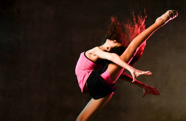 Jeune fille mince athlétique gymnaste danseuse sauter jusqu'à étirement faire des exercices de gymnastique en studio sur le mur sombre — Photo
