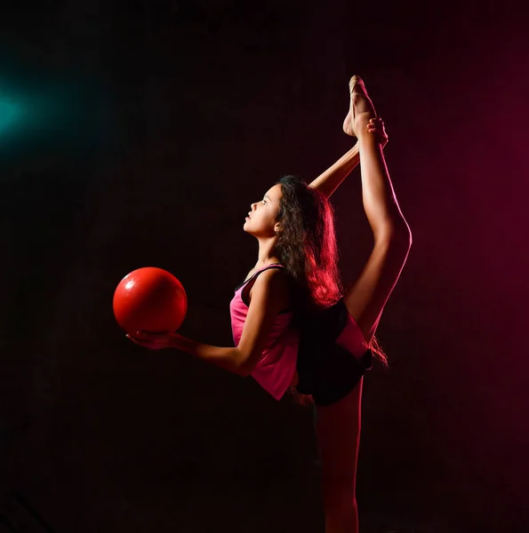 Young slim athletic girl gymnast doing gymnastic exercises with red ball and stretching in studio on dark wall — Stock Photo, Image