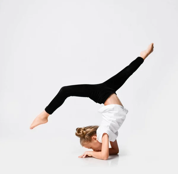 Slim athletic kid girl is doing gymnastic acrobatics exercises, stretching with her legs up on white — Stock Photo, Image