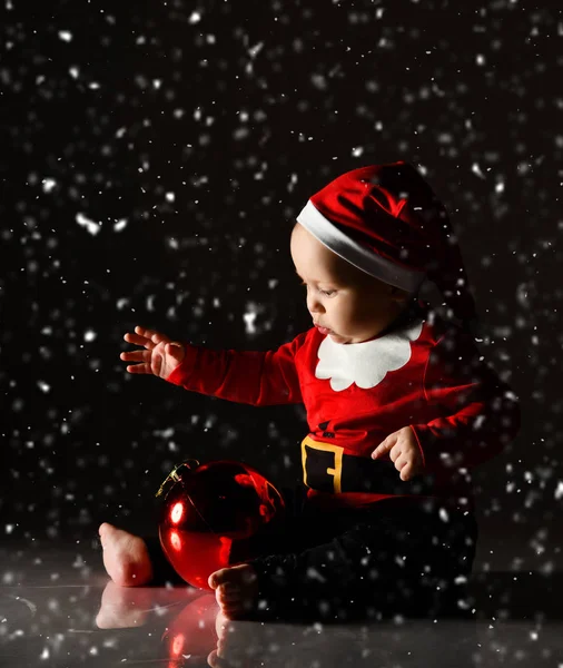 Dítě chlapeček batole sedí v kostýmu Santa Claus, čepice pod sněhem hraje s velkou červenou vánoční stromeček míč — Stock fotografie