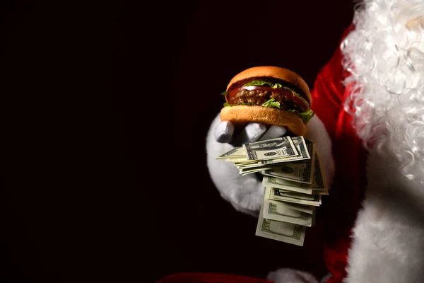 Babbo Natale ha un mucchio di soldi in una mano e un panino con doppio hamburger di manzo. Concetto fast food — Foto Stock