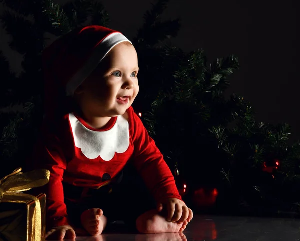 Kırmızı Noel Baba kostümlü ve şapkalı, yalınayak gülümseyen bebek bebek, yeni yıl hediyesi ile köknar arasında oturuyor. — Stok fotoğraf