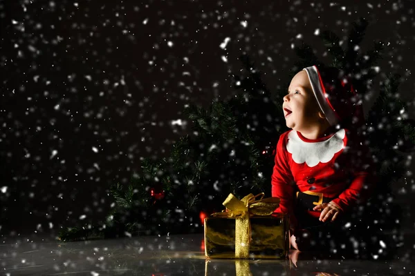 Petit garçon riant en costume de Père Noël claus est assis avec boîte cadeau sur le fond de l'arbre de Noël regardant la neige tomber — Photo