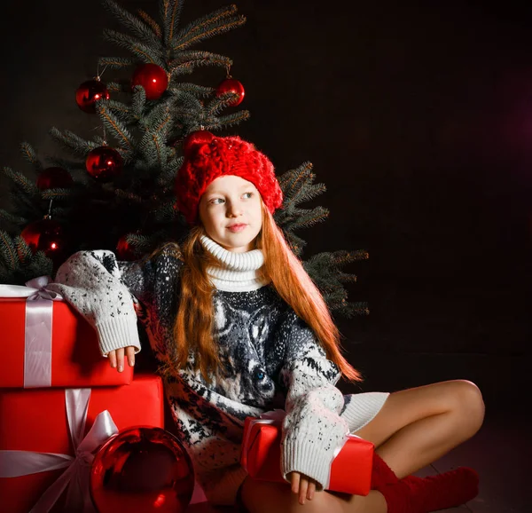 お祝いに座っている若い女の子は赤い冬の帽子でクリスマスツリーを飾り、ギフトボックスを提示します。新年のクリスマス — ストック写真