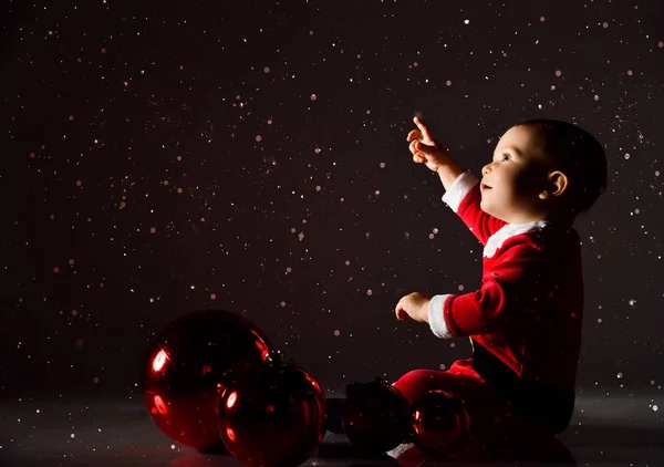 Ευτυχισμένο μικρό παιδί με κοστούμι Άγιος Βασίλης κάθεται στο πάτωμα σε μπάλες χριστουγεννιάτικου δέντρου που δείχνουν προς τα πάνω κάτω από το χιόνι — Φωτογραφία Αρχείου
