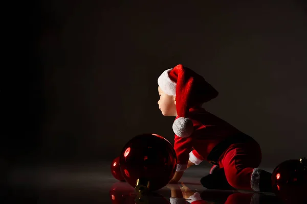 Μικρό παιδί σε κόκκινο κοστούμι Σάντα και καπέλο σέρνεται σε όλα τα τέσσερα μεταξύ χριστουγεννιάτικο δέντρο μπάλες διακόσμηση στο σκοτάδι — Φωτογραφία Αρχείου