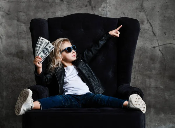 Zelfverzekerde rijke kind meisje zit in luxe fauteuil met bundel dollars cash kijken wijzend op de bovenste hoek — Stockfoto