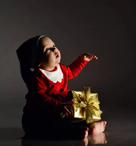 Βρέφος αγόρι νήπιο σε κοστούμι Santa Claus κάθεται με το νέο έτος δώρο κουτί παρουσιάζουν κρατώντας το χέρι επάνω, χαστούκια, ψάχνει — Φωτογραφία Αρχείου