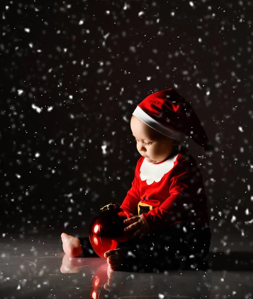 Kojenec chlapeček batole v Santa Claus kostým a čepice sedí na ledu pod sněhem hraje s vánoční stromeček míč — Stock fotografie
