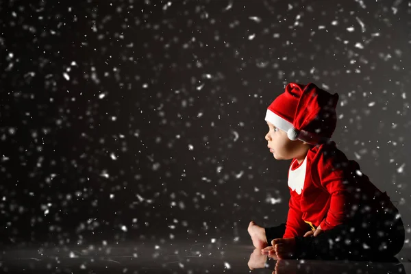 Βρέφος μικρό αγόρι νήπιο σε κόκκινο καπέλο Χριστούγεννα και το νέο έτος κοστούμι κάθεται στον πάγο κάτω από το χιόνι κοιτάζοντας φως ray — Φωτογραφία Αρχείου