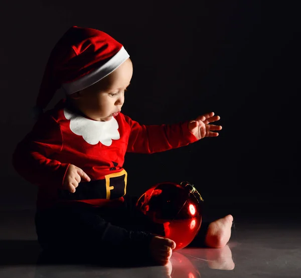 Παιδί μικρό αγόρι στο κοστούμι Santa Claus απασχολείται παίζοντας με ένα κόκκινο χριστουγεννιάτικο δέντρο μπάλα ελάτη διακόσμηση στο σκοτάδι — Φωτογραφία Αρχείου