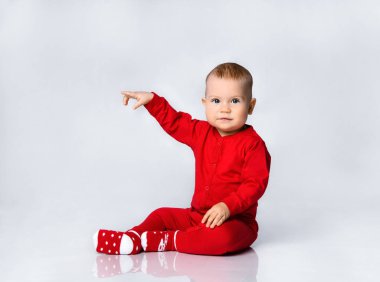Kırmızı bebek tulumlu küçük çocuk oturuyor ve bize bakıyor. Parmağını gri tüm vücuduna doğrultuyor.