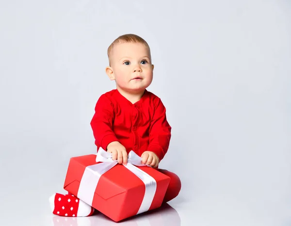 Niño pequeño en traje de bebé rojo está sentado con el regalo de Navidad de año nuevo, mirando hacia arriba pidiendo tratando de desatar — Foto de Stock