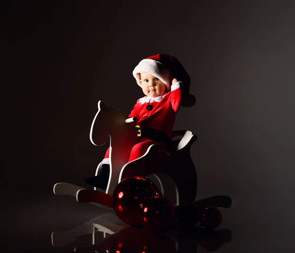 Niemowlę chłopiec maluch w czerwonym stroju Świętego Mikołaja jest jazda jego drewniany koń dla dzieci, trzymając jego Boże Narodzenie czapka — Zdjęcie stockowe