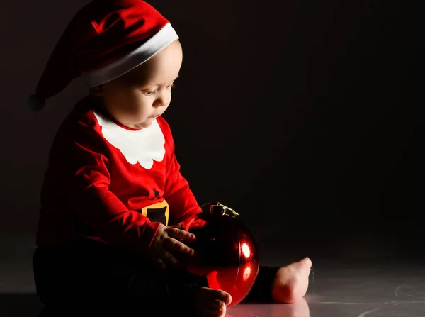 Noel Baba kostümlü bebek bebek, koyu renk yansımaya dokunan kırmızı Noel ağacı balosu süslemesiyle oynuyor. — Stok fotoğraf