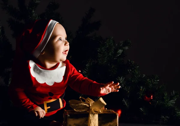 Šťastné dítě chlapeček v červeném kostýmu Santa Claus a čepice sedí a dívá se nahoru bude mít zlatý dárkový box — Stock fotografie