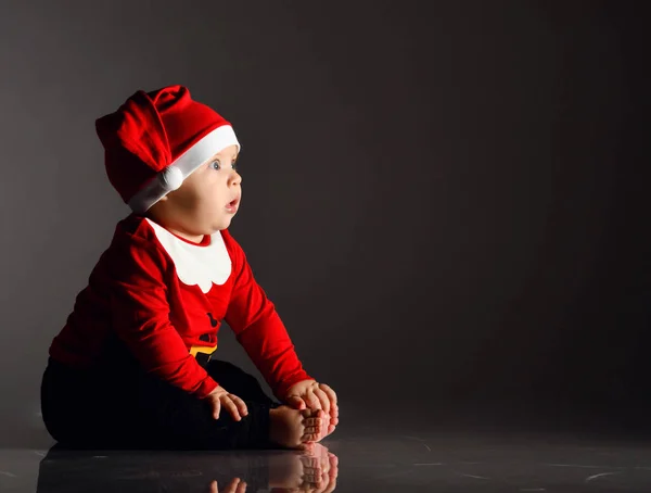 Zaskoczony lub przestraszony niemowlę chłopiec maluch siedzi w czerwony Święty Mikołaj kostium na lodzie na ciemno patrząc na przestrzeń kopiowania — Zdjęcie stockowe