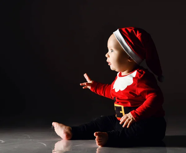 Kırmızı Noel Baba kostümü içinde büyülenmiş bebek bebek, buzun üzerinde oturmuş fotokopi çekilen yere dikkatlice bakıyor. — Stok fotoğraf