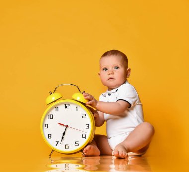 Infant baby boy toddler is occupied with sitting at big yellow alarm clock playing showing on yellow background clipart