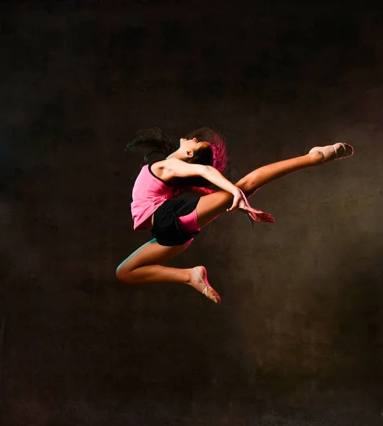 Junge schlanke, athletische Turnerin, die im Studio an dunkler Wand hochspringt und Dehnübungen macht — Stockfoto