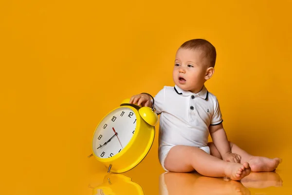 Uykulu bebek oğlan çocuğu elinde büyük sarı alarmlı bir saatle oturuyor. — Stok fotoğraf
