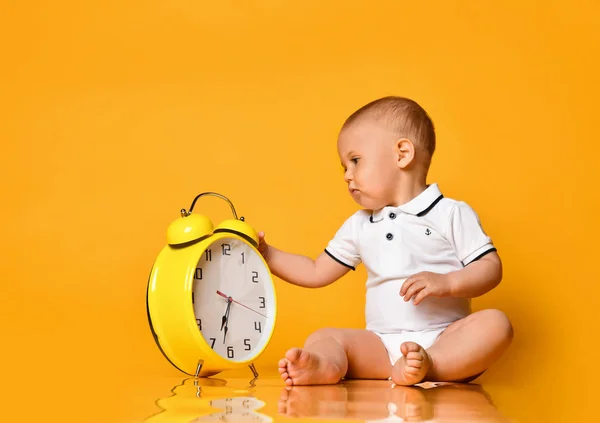 男婴蹒跚学步的婴孩被一只黄色的大闹钟占据着，它在黄色的背景下嬉戏探索 — 图库照片