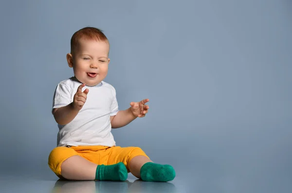 Infant baby boy toddler in yellow pant and white t-shirt is sitting on the floor laughing, sticking out his tongue — Stock Photo, Image