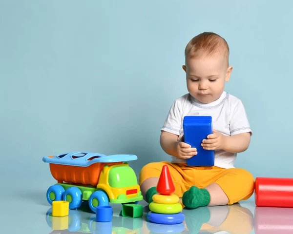 Infant baby boy toddler in yellow pants and white t-shirt is sitting on the floor surrounded by colorful toys — Stock Photo, Image