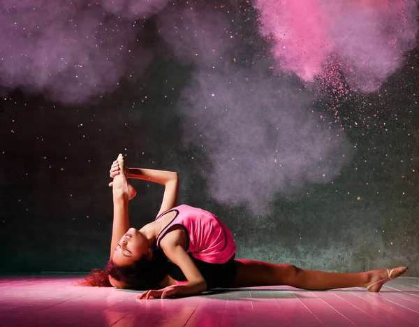 Young girl gymnast is doing gymnastic exercises on the floor does stretching with one her leg bent up in smoke splashes — Stock Photo, Image