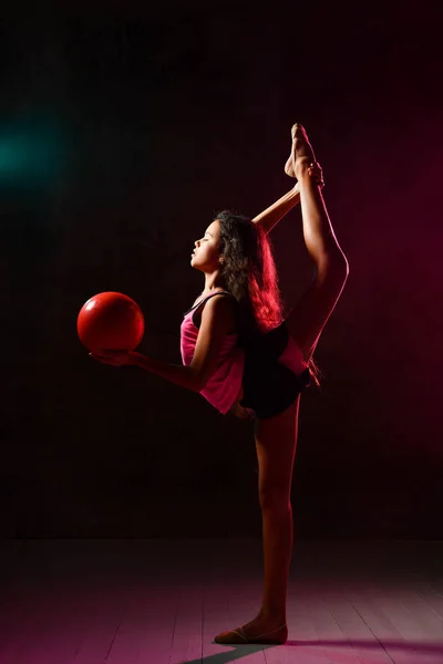 Молодая стройная атлетка гимнастка делает гимнастические упражнения с красным мячом и растягивается в студии на темной стене — стоковое фото