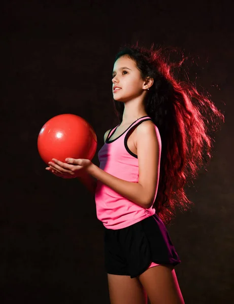 Young slim athletic girl gymnast doing gymnastic exercises with red ball and stretching in studio on dark wall — Stock Photo, Image