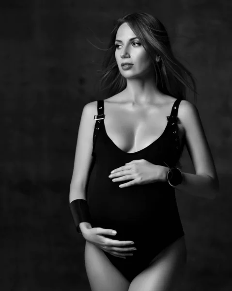 Jeune belle femme enceinte mince aux cheveux longs en corps noir et accessoires debout, embrassant le ventre en studio — Photo