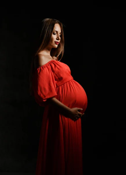 Νεαρή όμορφη έγκυος γυναίκα με κόκκινο φόρεμα στέκεται, αγκαλιάζει την κοιλιά και χαμογελά — Φωτογραφία Αρχείου