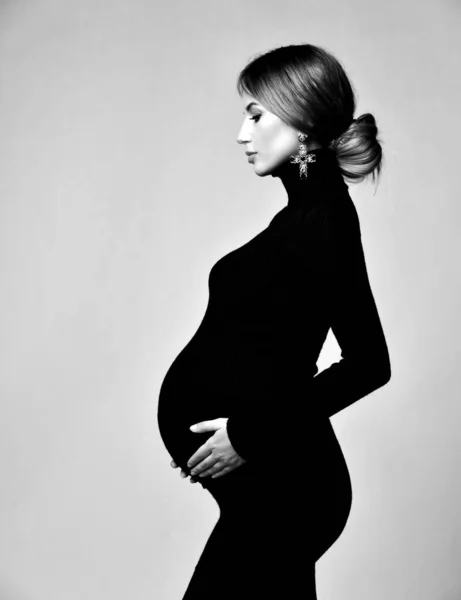 Young beautiful pregnant woman in black dress and massive earrings standing and looking down — 스톡 사진